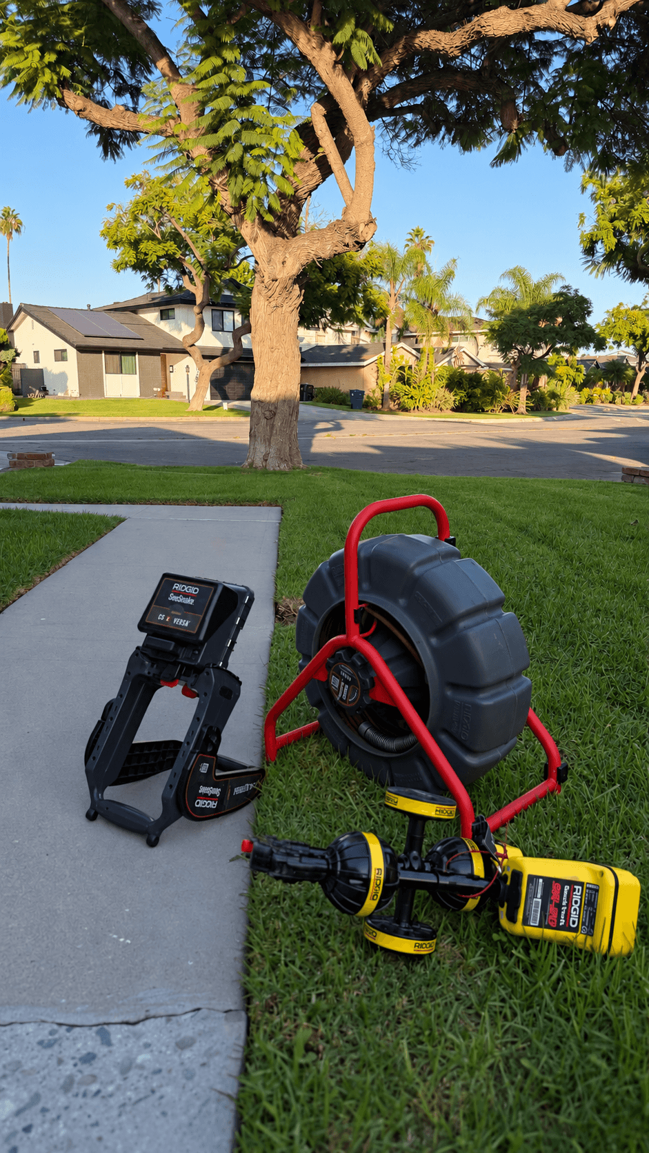 RIDGID plumbing inspection tools on a green lawn in a sunny suburban residential neighborhood.