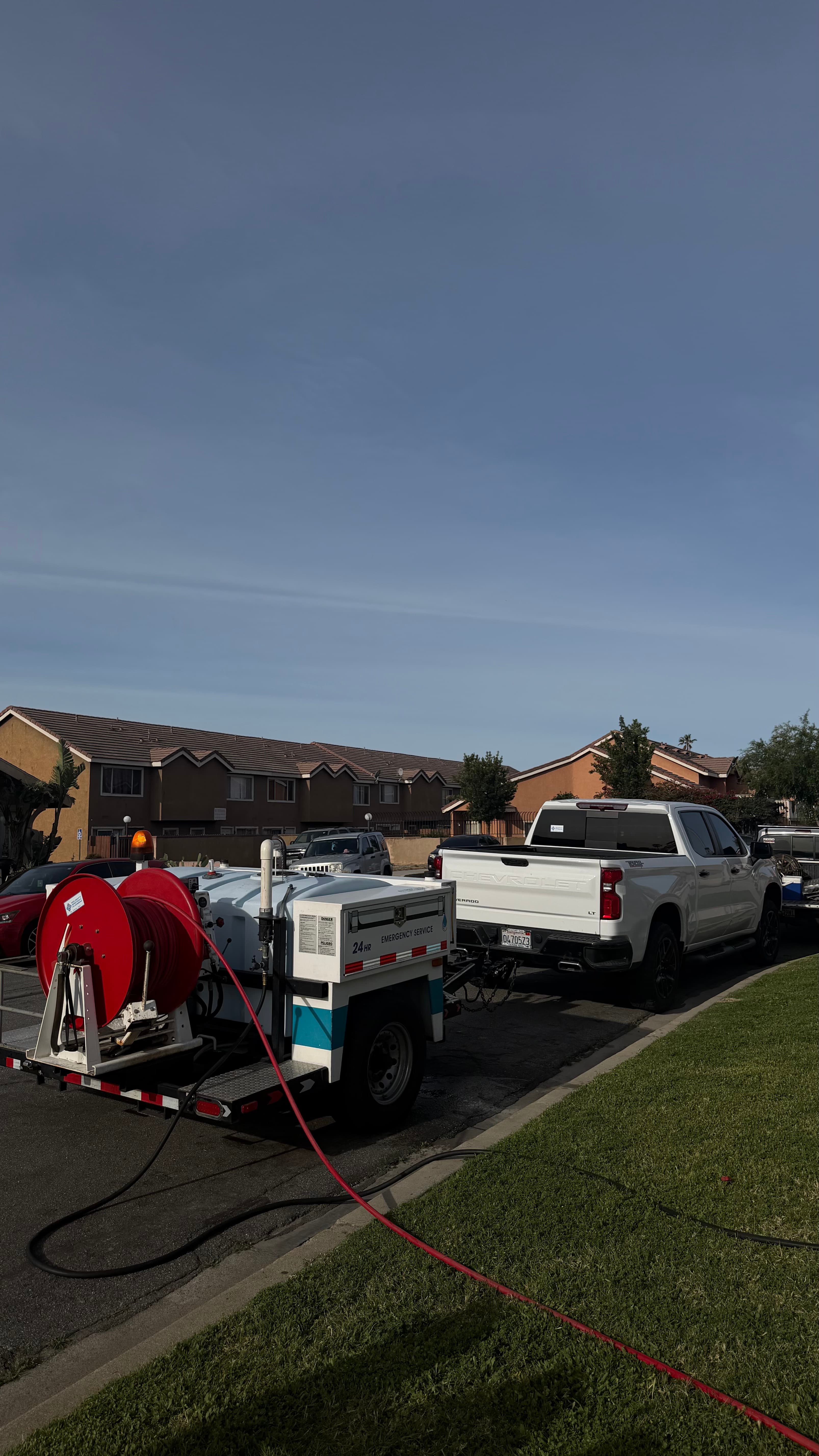 White Chevrolet pickup towing an emergency service trailer with a red hose extended across grass.