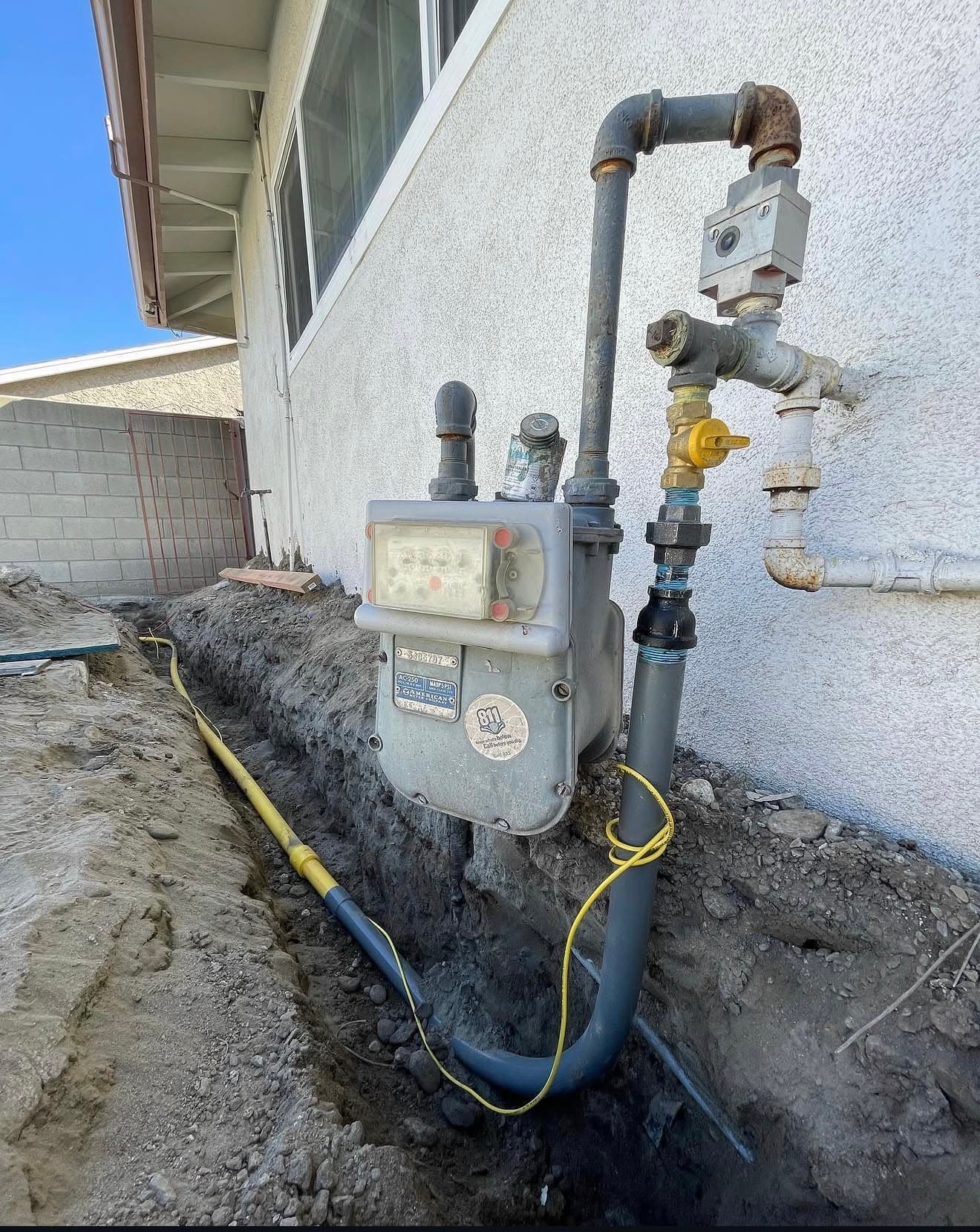 Residential gas meter on a house wall with a yellow pipe in a trench.